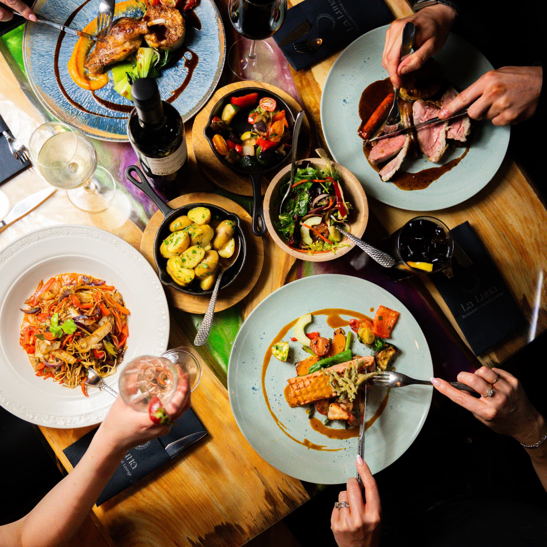 Overhead view of a shared La Luna table spread with steak, duck, noodles, salmon, sides and wine